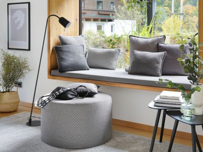 Ein gemütlicher Wohnbereich mit einer Fensterbank, Kissen, einem runden Hocker und einer Stehlampe. Pflanzen und Bücher ergänzen die Einrichtung.
